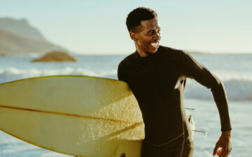 Smiling african male running out of the ocean after water surfing. Happy young man with surfboard on the beach enjoying holidays.